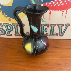 May include: A black ceramic pitcher with a handle and a flared spout. The pitcher has colorful abstract accents in yellow, teal, and red. The word "Ecolon" is written on the side.