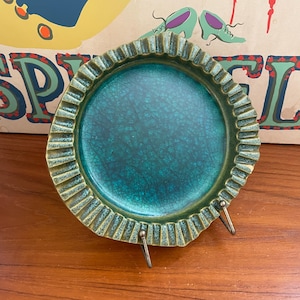 May include: A teal and green ceramic ashtray with a textured, ruffled edge. The ashtray has a crackle glaze finish and is supported by a metal stand. The background features a vintage poster with text and illustrations.