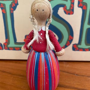 May include: A wooden doll with blonde braids, wearing a red dress with blue and orange stripes. The doll has a painted face with black eyes and a white collar.