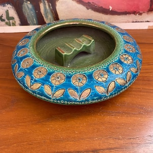 May include: A round ceramic ashtray featuring a vibrant turquoise blue glaze with a repeating gold-toned floral pattern. The interior is finished in a deep olive green glaze and includes a central bridge-shaped rest for cigarettes.