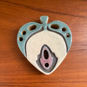 May include: A heart-shaped ceramic wall hanging with a white center and a blue and brown design. The center has a small, round hole and a larger, oval hole.