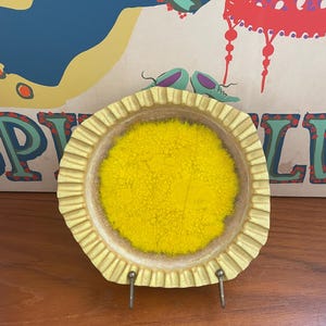 May include: A round, yellow and beige ceramic dish with a textured yellow center and a ruffled edge. The dish is displayed on a metal stand against a colorful background with text.
