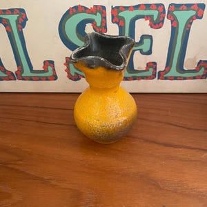 May include: A small, yellow vase with a dark gray, star-shaped rim. The vase has a rounded body and a textured surface. The background features a colorful design with the letters "SEL".