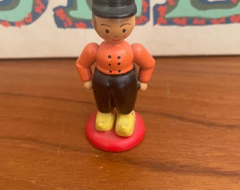 May include: A small, wooden figurine of a person wearing a black hat, orange jacket with black buttons, and black pants. The figurine stands on a red base and wears yellow shoes. The background is a wooden surface.