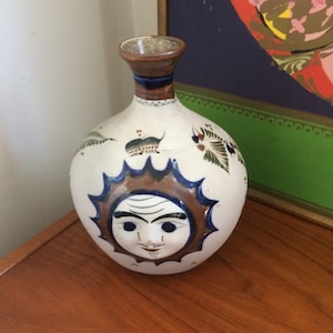 May include: A white ceramic vase with a round body and a narrow neck. The vase features a sun face design in brown and blue, with additional bird and floral motifs. The vase is displayed on a wooden surface.
