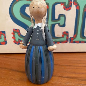 May include: A wooden doll with blonde hair in pigtails, wearing a black dress with white buttons and a blue and black striped skirt.