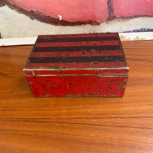 May include: A small, rectangular, red metal box with black stripes on the top. The box is slightly worn and has a rustic look.