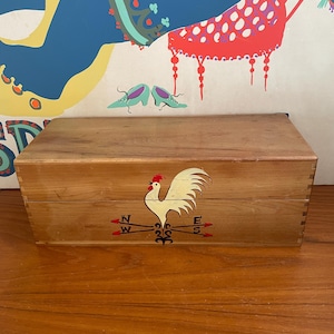 May include: A wooden box with a rooster weathervane design on the front. The weathervane features a white rooster with a red comb and the cardinal directions. The box is made of light-colored wood.