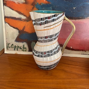 May include: A white ceramic pitcher with a black and gold geometric design. The pitcher has a turquoise blue glaze on the inside and a gold handle.