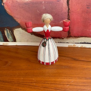May include: A wooden figurine of a woman in a traditional dress, holding two red candle holders. The figurine has a blonde hair and a red and white dress with black stripes. The base is red and white striped. The background is a painting with blue and pink colors.