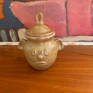 May include: A small, brown ceramic jar with a face sculpted on the front. The jar has a lid with a loop handle and two small handles on the sides.