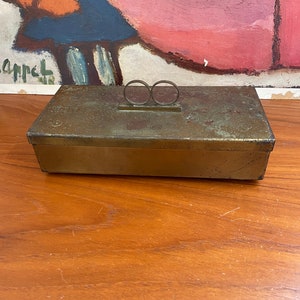 May include: A vintage brass box with a hinged lid and a decorative ring handle. The box has a worn, aged patina and is decorated with a repeating pattern of small circles.
