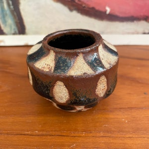 May include: A small, brown ceramic vase with a geometric pattern of white and black accents. The vase has a rounded body and a slightly flared opening.