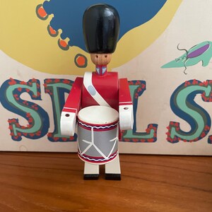 May include: A wooden toy soldier drummer in a red uniform with a black hat and a white strap. The drum is gray with a white rim and a red and blue design. The toy is standing in front of a colorful background.