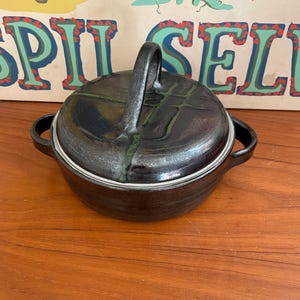 May include: A dark brown ceramic casserole dish with a lid and a handle. The lid has a green and black design. The dish has a silver rim and two side handles. The background includes a vintage poster with the words "SPIT SEL".