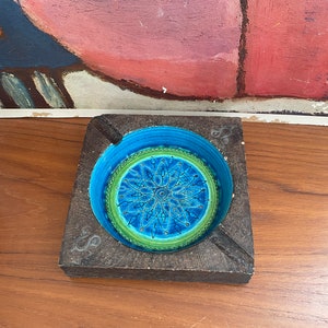 May include: A square wooden ashtray with a blue and green ceramic inset featuring a geometric design. The ashtray has a carved design on the wood.