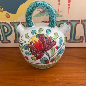May include: A white ceramic pitcher with a teal handle and spout. The pitcher is decorated with a large red and yellow floral design, green leaves, and small red dots. The handle has a textured appearance.