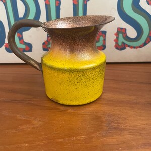 May include: A small, vintage creamer with a yellow body and a brown top. The creamer has a handle and a spout. The handle and rim are a darker brown color. The creamer is likely made of metal.