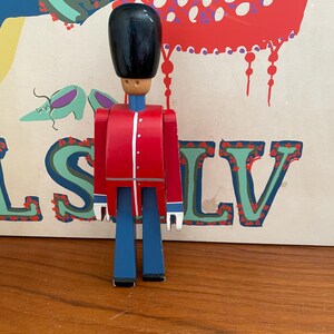 May include: A wooden toy soldier in a red jacket with white trim, blue pants, and a black hat. The toy soldier has a light brown head and is standing in front of a colorful background.