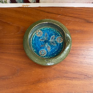 May include: A small, green ceramic ashtray with a blue and gold floral design. The ashtray has a slightly raised rim and a shallow well.