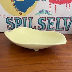 May include: A pale yellow, leaf-shaped ceramic dish. The dish has a smooth surface and a small, rounded foot. The background features a vintage poster with the text "SPIL SELV".