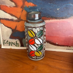 May include: A ceramic vase with a gray background and a geometric pattern of green, yellow, orange, and white leaves. The vase is about 10 inches tall.
