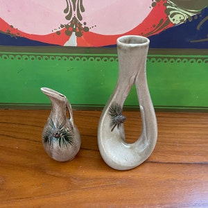 May include: Two ceramic vases with a pine cone design. The larger vase is a light brown color and has a unique, curved shape. The smaller vase is a darker brown color and has a more traditional shape. Both vases have a green and brown pine cone design.
