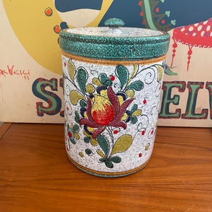 May include: A white ceramic canister with a green lid and a floral design in red, yellow, green, and blue. The canister has a speckled texture.
