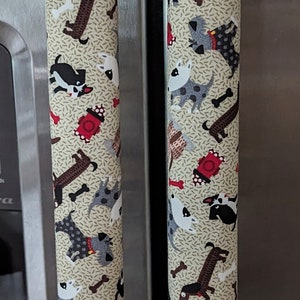 May include: Two stainless steel refrigerator handles with beige fabric covers. The fabric covers feature a repeating pattern of dogs in various poses and colors, including white, black, brown, and gray. The dogs are surrounded by small red fire hydrants and white bone shapes.