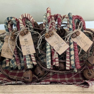 May include: Handmade fabric candy canes with tags that read "Candy Cane Co" and "Believe". The candy canes are red, white, and green plaid. They are tied with twine and have small bells attached. The arrangement sits on a plaid fabric.