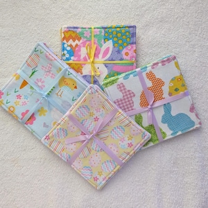 May include: Four square fabric coasters with Easter-themed patterns, including eggs, bunnies, and flowers. Each set is tied with a ribbon in various colors, such as yellow, pink, and purple. The coasters are on a white surface.