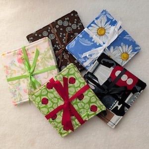 May include: A collection of six square fabric coasters, each with a unique pattern and tied with a ribbon. Designs include floral, ladybug, and geometric prints. Ribbons are in various colors, including green, red, and brown.