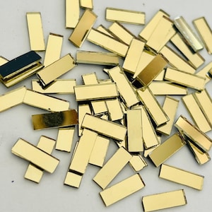 May include: A pile of small, rectangular gold mirror tiles. The tiles are scattered on a white surface.