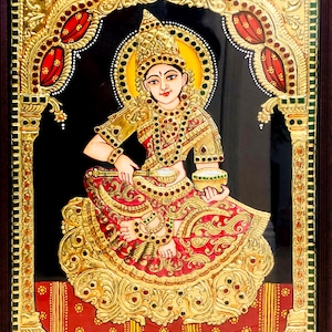 Puede incluir: Pintura enmarcada y ornamentada de una figura sentada con vestimenta tradicional india. La obra presenta colores dorados, rojos y negros con detalles intrincados. La figura está adornada con joyas y sostiene cuencos, sobre un fondo oscuro.