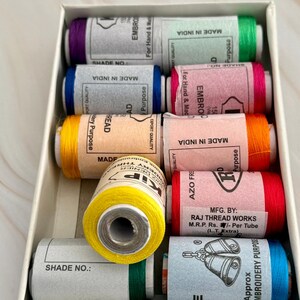 May include: A rectangular box filled with spools of colorful embroidery thread. The spools are wrapped in paper labels with text and are in shades of purple, green, blue, pink, orange, yellow, and red. The box is white.
