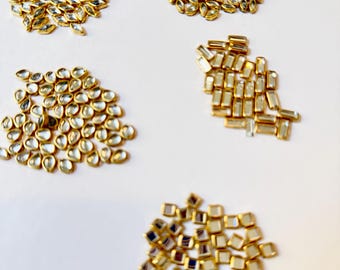 Puede incluir: Una colección de pequeños adornos decorativos para manualidades con monturas de metal dorado e inserciones de vidrio transparente y reflectante. Las piezas están dispuestas en cinco grupos distintos, mostrando varias formas geométricas, incluyendo diseños de marquesa, lágrima, rectangulares y cuadrados.