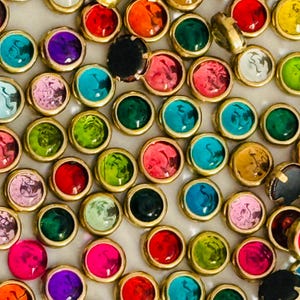 May include: A collection of colorful, round cabochons with gold-colored metal settings. The cabochons are in various colors, including pink, blue, green, orange, and purple. Some have a clear, glass-like appearance, while others are opaque.