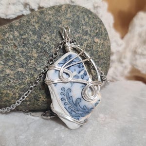 May include: A handcrafted pendant necklace featuring a piece of blue and white patterned ceramic, wire-wrapped in silver-colored metal. The pendant hangs from a delicate silver chain. The ceramic has a floral design.