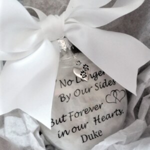 May include: White glass ornament with a silver paw print charm. The ornament has the text "No Longer By Our Side But Forever in Our Hearts. Duke" printed on it.