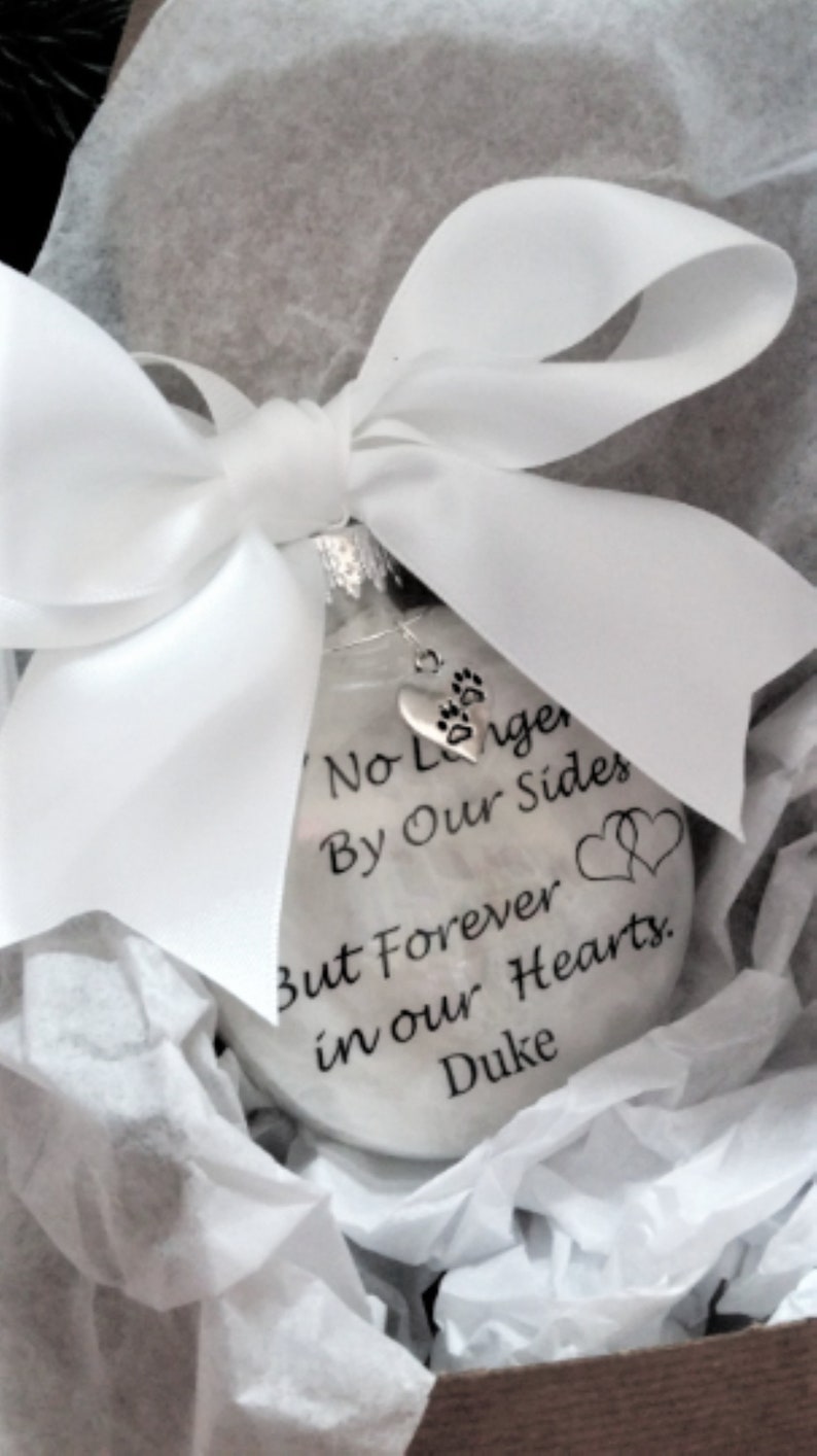 May include: White glass ornament with a silver paw print charm. The ornament has the text "No Longer By Our Side But Forever in Our Hearts. Duke" printed on it.