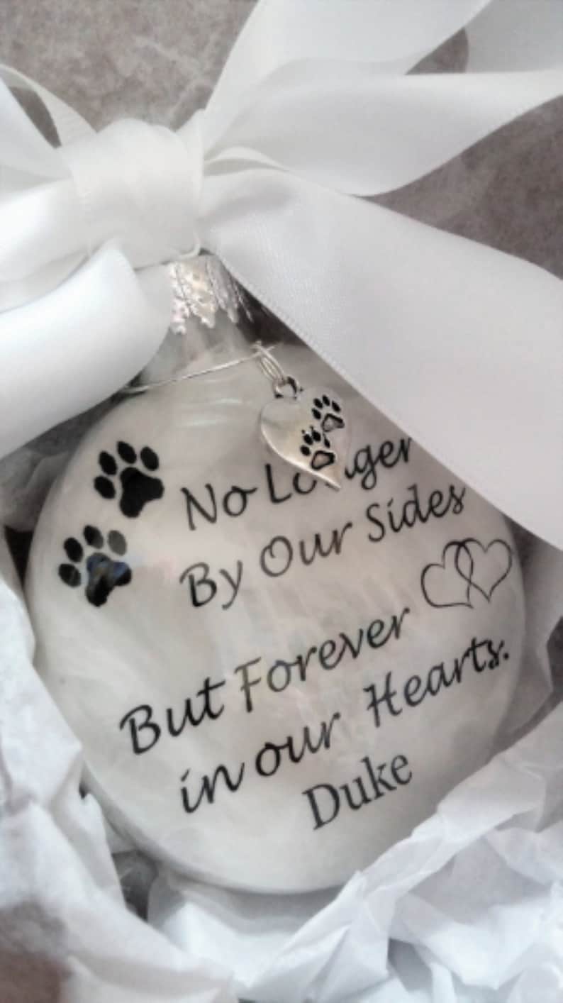May include: White glass ornament with a black paw print design and the text "No Longer By Our Sides But Forever in our Hearts. Duke".