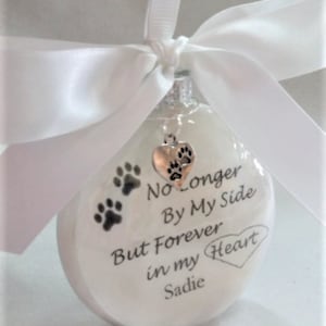 May include: White glass ornament with a silver heart charm and paw prints. The ornament reads "No Longer By My Side But Forever in my Heart Sadie".