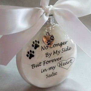May include: White glass ornament with a black paw print design and the text "No Longer By My Side But Forever in my Heart Sadie". A silver charm with a paw print heart hangs from the ornament.