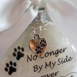 May include: White glass ornament with a silver heart charm with paw prints. The ornament has black paw prints and the text "No Longer By My Side Forever In My Heart".
