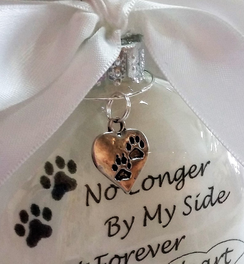 May include: White glass ornament with a silver heart charm with paw prints. The ornament has black paw prints and the text "No Longer By My Side Forever In My Heart".