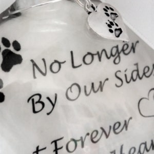 May include: White glass ornament with a silver paw print charm. The ornament has black text that reads "No Longer By Our Side But Forever in our Hearts Duke".