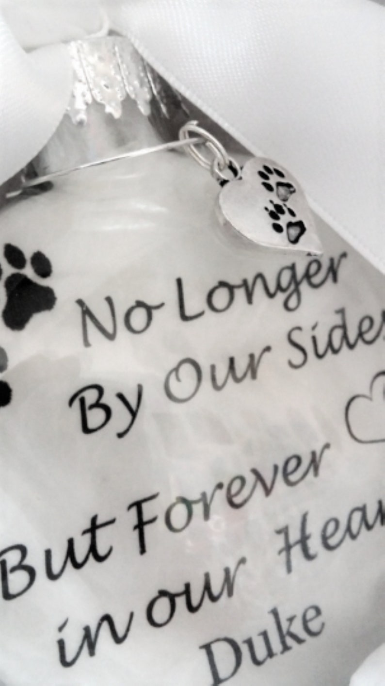 May include: White glass ornament with a silver paw print charm. The ornament has black text that reads "No Longer By Our Side But Forever in our Hearts Duke".