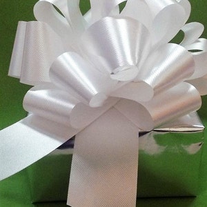 K&ouml;nnte beinhalten: Eine wei&szlig;e Satin Schleife, die auf einem silbernen Geschenkbox gebunden ist. Die Schleife ist gro&szlig; und flauschig, mit mehreren Schleifen und B&auml;ndern. Die Geschenkbox ist in silbernes Papier eingewickelt und hat einen gr&uuml;nen Hintergrund.