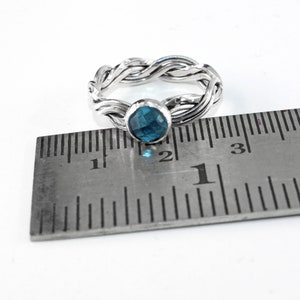 May include: A silver ring with a braided band and a blue gemstone. The ring is measured against a ruler, showing it is slightly over 2.5 cm in diameter.
