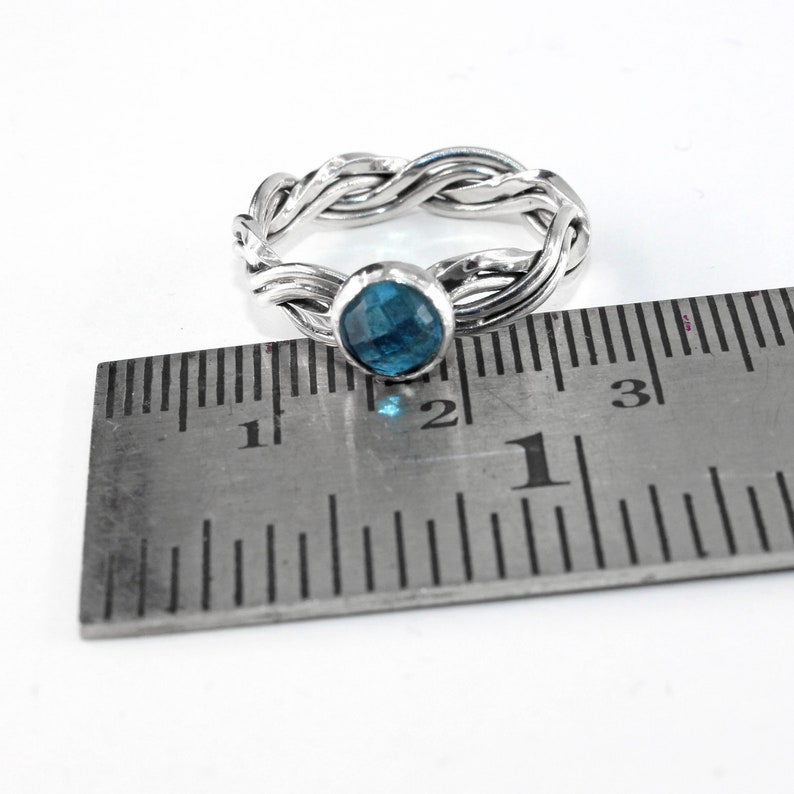 May include: A silver ring with a braided band and a blue gemstone. The ring is measured against a ruler, showing it is slightly over 2.5 cm in diameter.